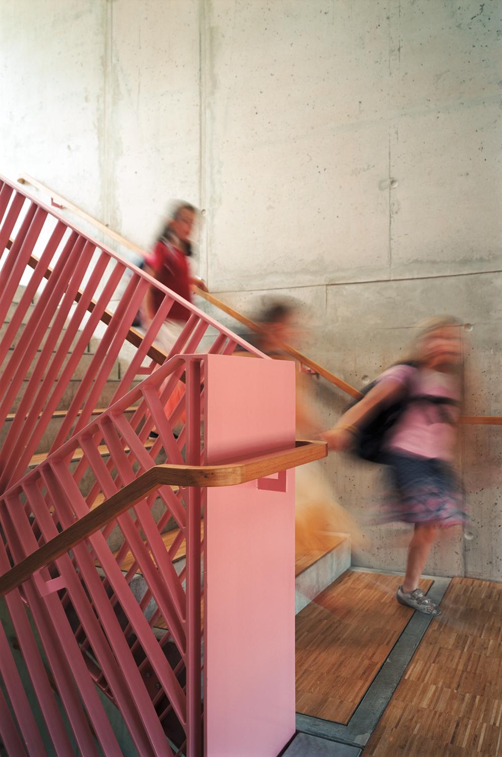 Sichtbetonoberflächen prägen die Wandgestaltung im Innern. In Kombination mit Bodenbelägen aus Holz und farbigen Akzenten entsteht eine angenehme, unprätentiöse Farbstimmung. Das Motiv des Geländers korrespondiert zudem mit der Umwehrung der Schulhofs auf dem Dach.