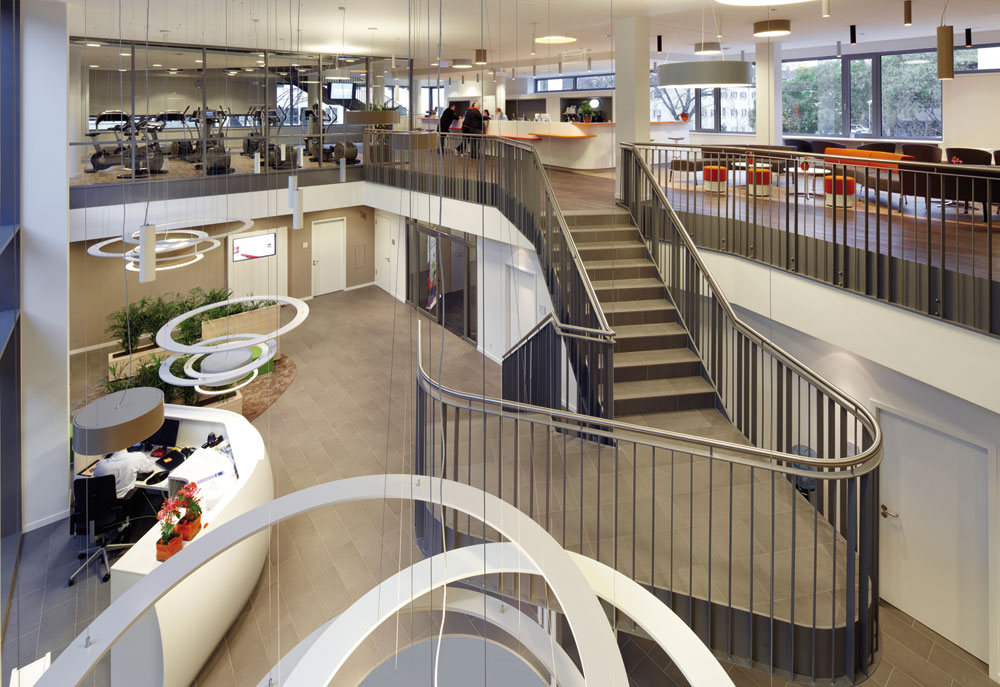 Über eine skulpturale Treppe gelangt man vom Foyer in den Empfangsbereich des Fitnesstudios und die Café-Bar.