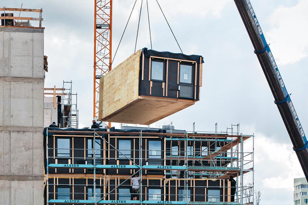Vorgefertigte Brettsperrholz-Module bzw. -Wandelemente sowie die Betonfertigteile der Flurkonstruktion wurden angeliefert und sofort an die entsprechende Stelle im Ge- bäude eingehoben. Eine Ausgleichsschicht aus Holzschwellen sorgt auf dem Betontisch über dem EG für eine präzise ebene Fläche zum Stapeln der Module.