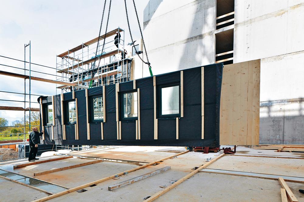 Vorgefertigte Brettsperrholz-Module bzw. -Wandelemente sowie die Betonfertigteile der Flurkonstruktion wurden angeliefert und sofort an die entsprechende Stelle im Ge- bäude eingehoben. Eine Ausgleichsschicht aus Holzschwellen sorgt auf dem Betontisch über dem EG für eine präzise ebene Fläche zum Stapeln der Module.
