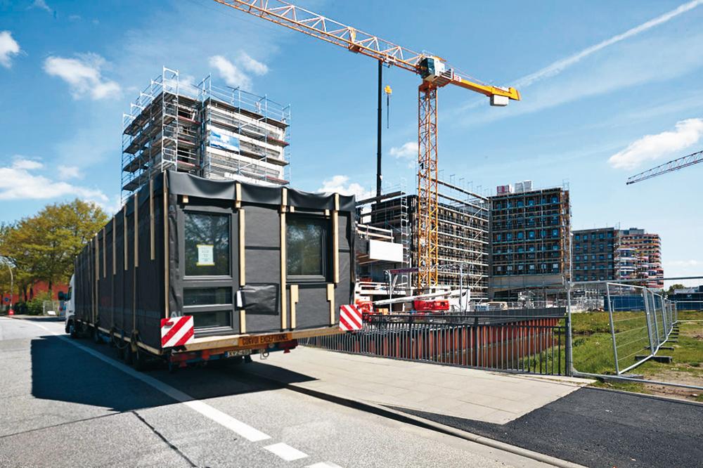 Vorgefertigte Brettsperrholz-Module bzw. -Wandelemente sowie die Betonfertigteile der Flurkonstruktion wurden angeliefert und sofort an die entsprechende Stelle im Ge- bäude eingehoben. Eine Ausgleichsschicht aus Holzschwellen sorgt auf dem Betontisch über dem EG für eine präzise ebene Fläche zum Stapeln der Module.