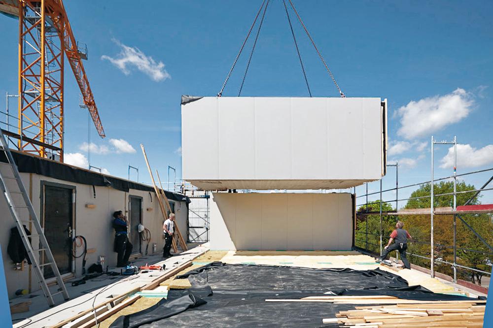 Vorgefertigte Brettsperrholz-Module bzw. -Wandelemente sowie die Betonfertigteile der Flurkonstruktion wurden angeliefert und sofort an die entsprechende Stelle im Ge- bäude eingehoben. Eine Ausgleichsschicht aus Holzschwellen sorgt auf dem Betontisch über dem EG für eine präzise ebene Fläche zum Stapeln der Module.