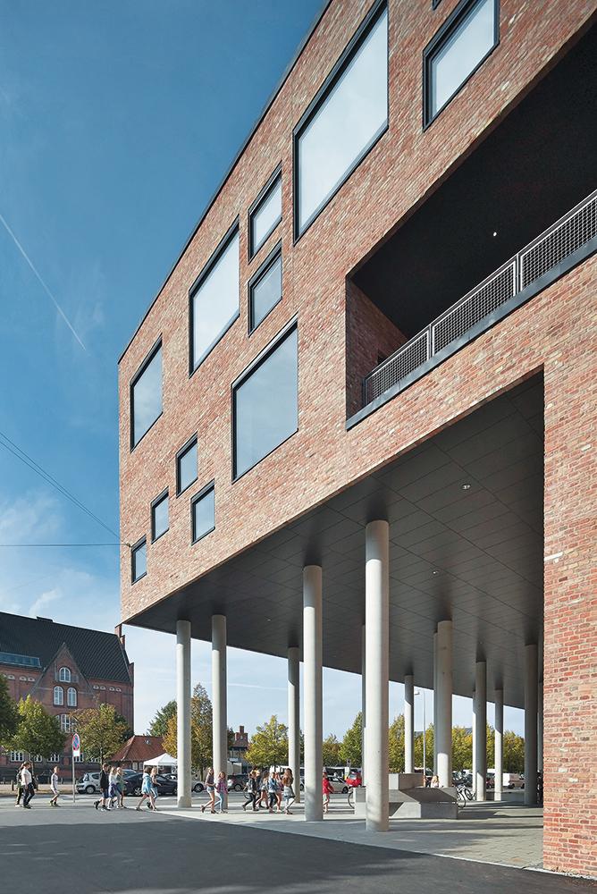 Der Eingang über Eck ist durch zweigeschossige Sichtbetonstützen markiert und bildet einen beliebten Treffpunkt. Vorplatz, Halle und Pausenhof sind öffentliche Räume, in denen sich die Wege
von Erwachsenen, Jugendlichen und Kindern aller Altersstufen kreuzen.