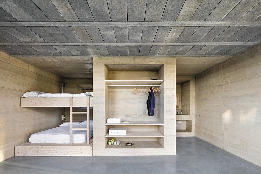 In einer Art Enfilade reihen sich die Wohn- und Schlafzimmer L-förmig an der Grundstücksgrenze auf. Die Trennwände sowie die Wandnischen für Regale und Waschbecken bestehen aus Stampfbeton.