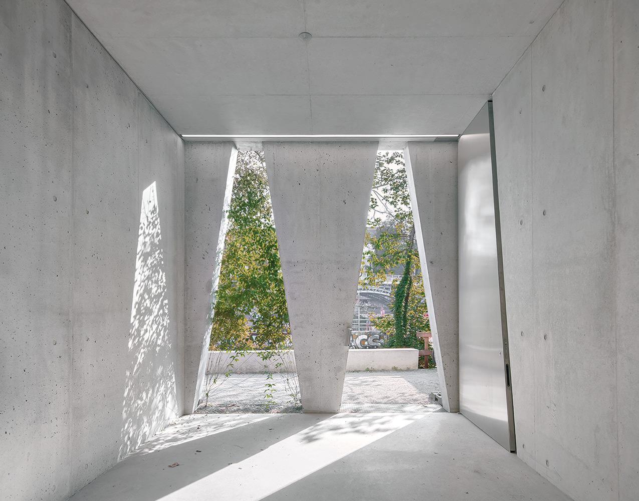 Das Foyer läuft parallel zum Flussufer über die gesamte Gebäudelänge. Der natürliche Sonnenschutz aus Kletterpflanzen, die an Stahlseilen empor ranken, malt Schattenbilder auf den grauen Sichtbeton.