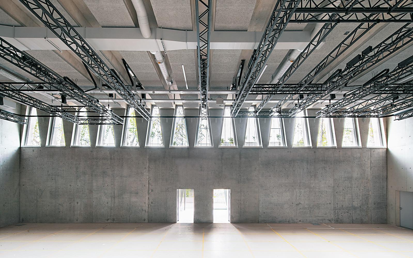 Das Foyer läuft parallel zum Flussufer über die gesamte Gebäudelänge. Der natürliche Sonnenschutz aus Kletterpflanzen, die an Stahlseilen empor ranken, malt Schattenbilder auf den grauen Sichtbeton.