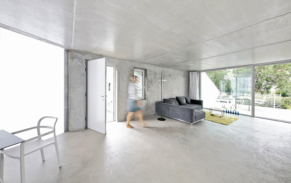 Ohne Vordach oder Windfang gelangt man direkt ins puristische eingerichtete Wohnzimmer. Dort öffnet sich der Raum über eine verglaste Schiebetür zum Balkon.