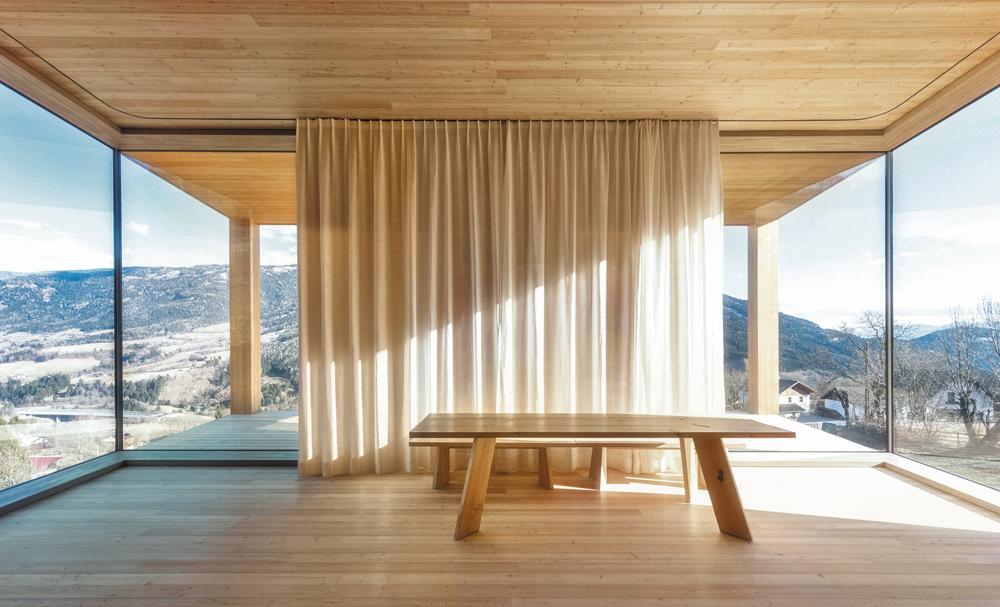 Mit großen Eckverglasungen öffnet sich der Wohnraum talwärts. Der Vorhang verbirgt eine geschlossene Fassadenpartie mit Regalen und Terrassentür.