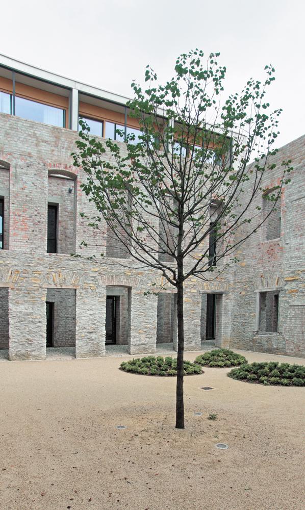 Wie eine Kulisse ist die Kreuzgangmauer freigestellt. Die dahinterliegende Bestandswand wurde zur Bürofassade mit verglasten Öffnungen ertüchtigt. Vom Kreuzgang aus ist der Neubau nur an der Aufstockung des Nordflügels abzulesen. Die übrigen Dachaufbauten sind im Duktus des Bestandsmauerwerks als Lochfassaden aus Ziegel ergänzt.