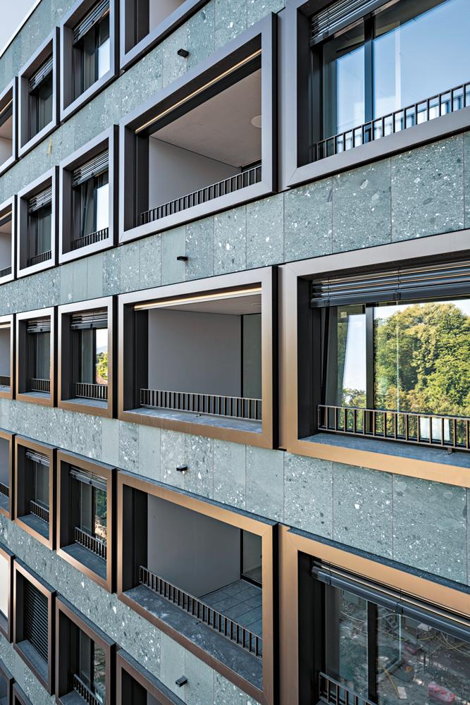 Die Fenster und Loggienöffnungen sind gleichermaßen mit breiten Aluminiumrahmen eingefasst. Die geschlossenen Fassadenflächen wurden mit Verde Salvan, einem grünen Gneiskonglomerat, verkleidet.