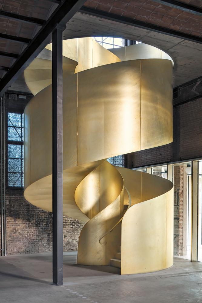 Die goldfarbene Treppenskulptur aus verzinktem Stahl bildet einen scharfen Kontrast zur historischen Textur des ehemaligen Industriegebäudes.