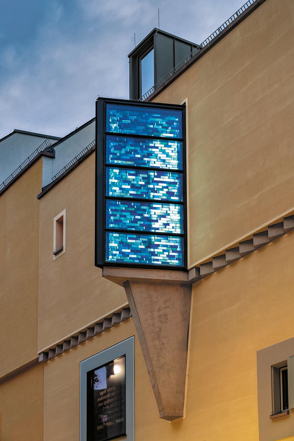 Die blaue Bleiverglasung hinter der äußeren Isolierglashülle taucht den Erkerraum tagsüber in blaues Licht. Nachts strahlt sie, elektrisch hinterleuchtet, in den Straßenraum hinaus.