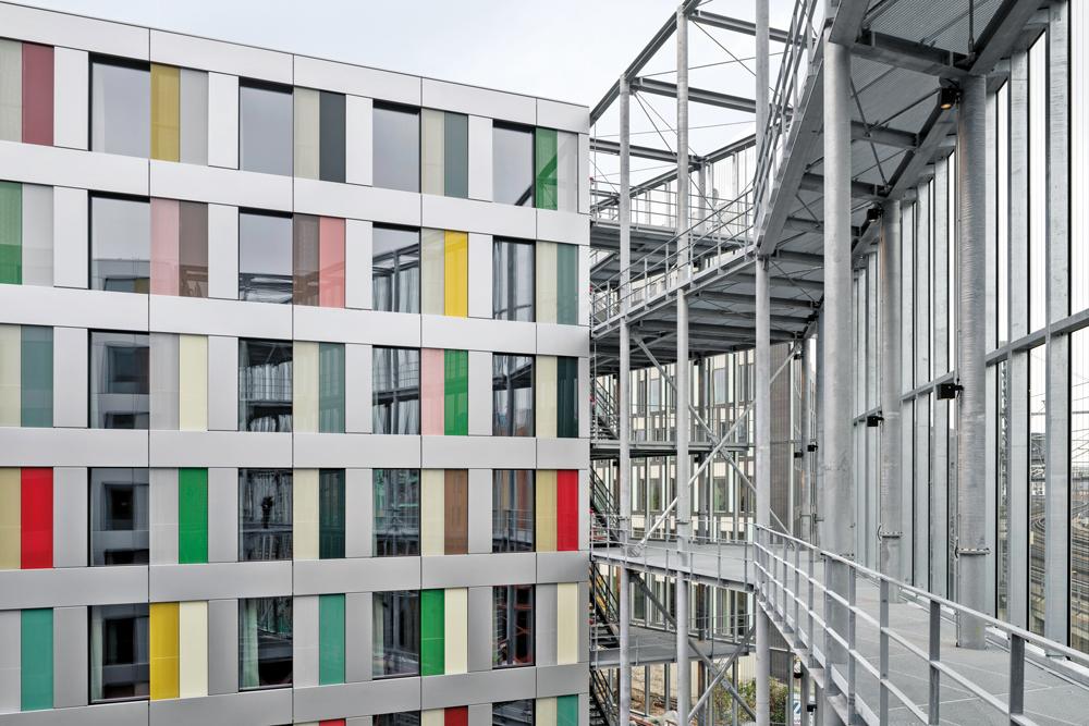 Auf der Nordseite verbindet ein offenes Stahltreppenhaus die Bürotrakte. Eine Glaswand schützt das Gebäude vor dem Lärm der angrenzenden Bahntrasse.