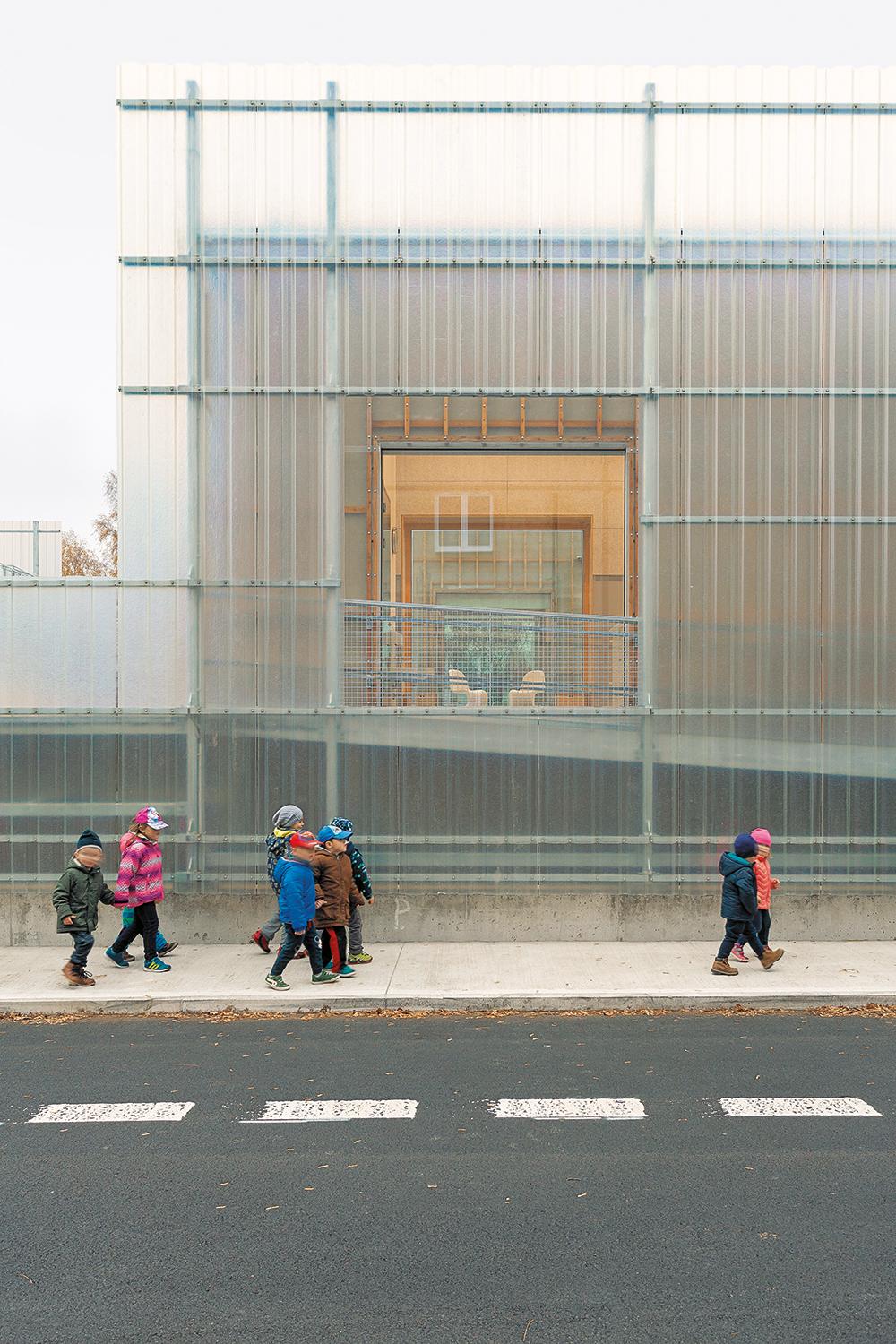 Hinter den gewellten Kunststoffplatten der äußeren Fassade erscheint schemenhaft der Massivbau des Kindergartens und das Stahlgerüst im Zwischenraum.