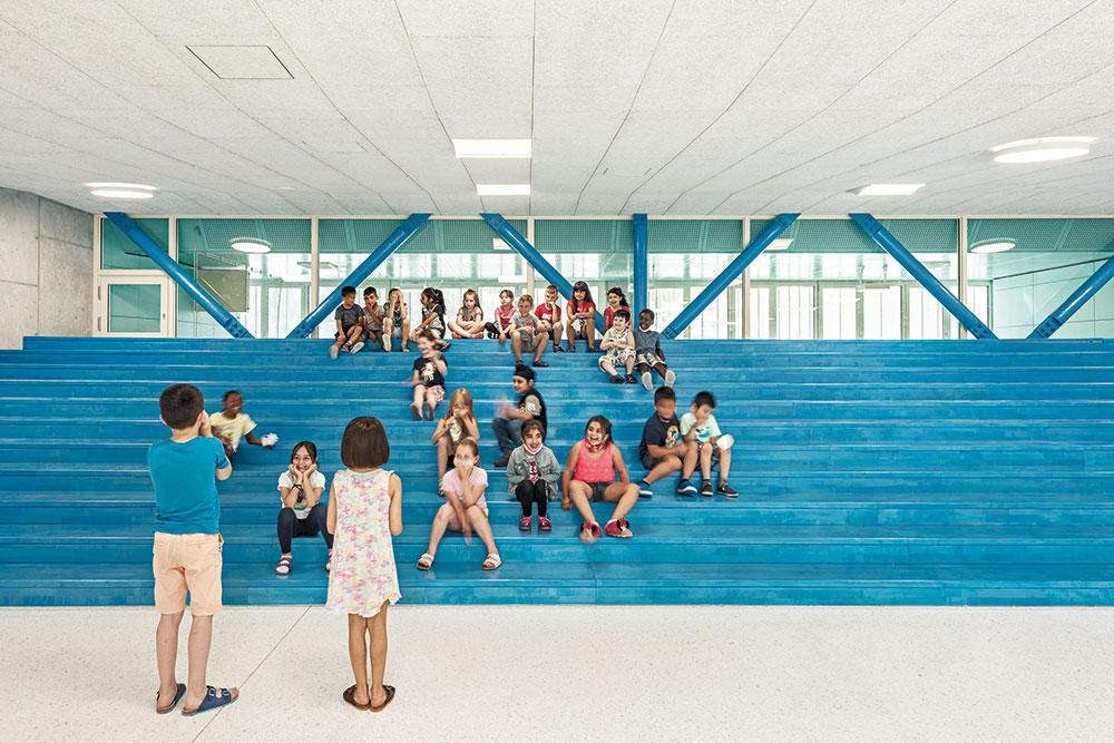 Begegnungsraum im Erdgeschoss: Die blauen Sitzstufen bieten zusätzliche Lernund Aufenthaltsbereiche. Die Verglasung zur Gymnastikhalle öffnet den Raum und holt Tageslicht herein.