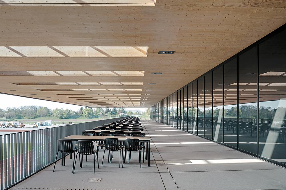 Die Cafeteria liegt zwischen den Sporthallen und der überdachten Terrasse. Die Zugstützen des auskragenden Holzdachs sind trotz der Brandschutzummantelung filigran.