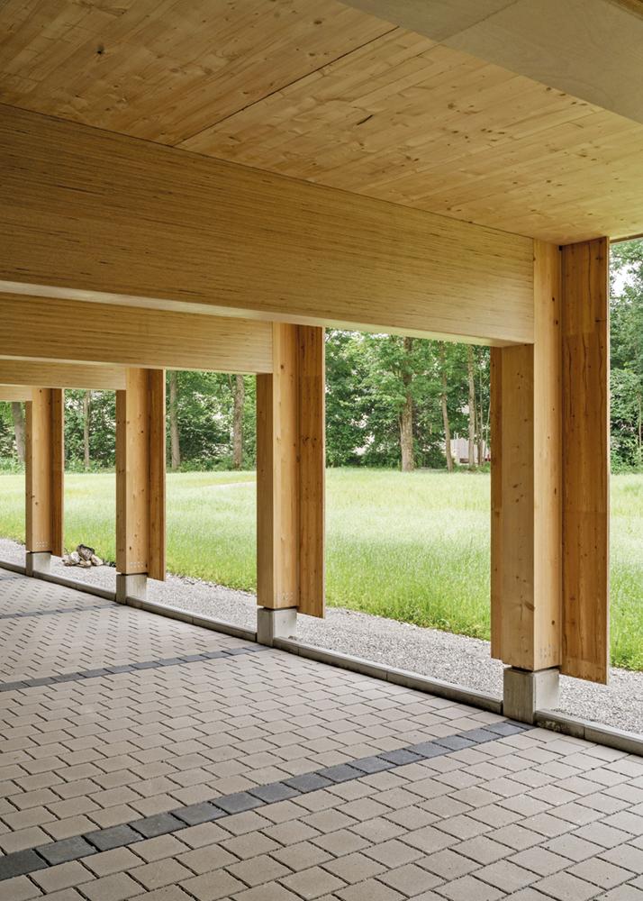 Über einem mit Betonpflaster belegten Parkplatz erhebt sich die zimmermannsmäßige Konstruktion. Träger und Stützen im Erdgeschoss sind aus Baubuche.
