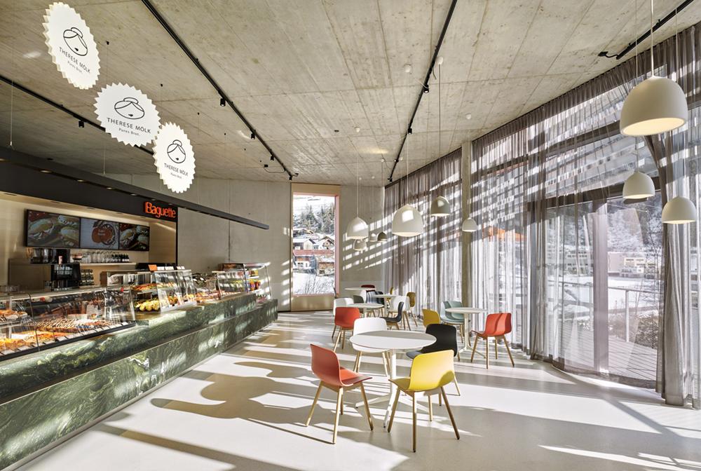 Hinter der Südfassade des Obergeschosses liegt ein BäckereiCafé. Hier weitet sich der Raum trichterartig zu einer schmalen Terrasse. Große Aluminiumlamellen spenden vor allem im Sommer Schatten.
