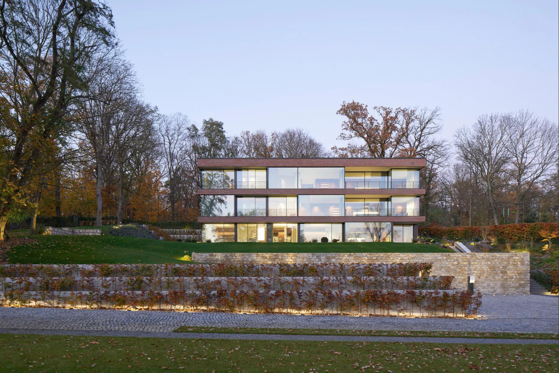 Durch die tiefen Rücksprünge und Freisitze in der Glasfassade verändert sich das Erscheinungsbild der Villa, abhängig von der gegenwärtigen Lichtstimmung und Tageszeit.