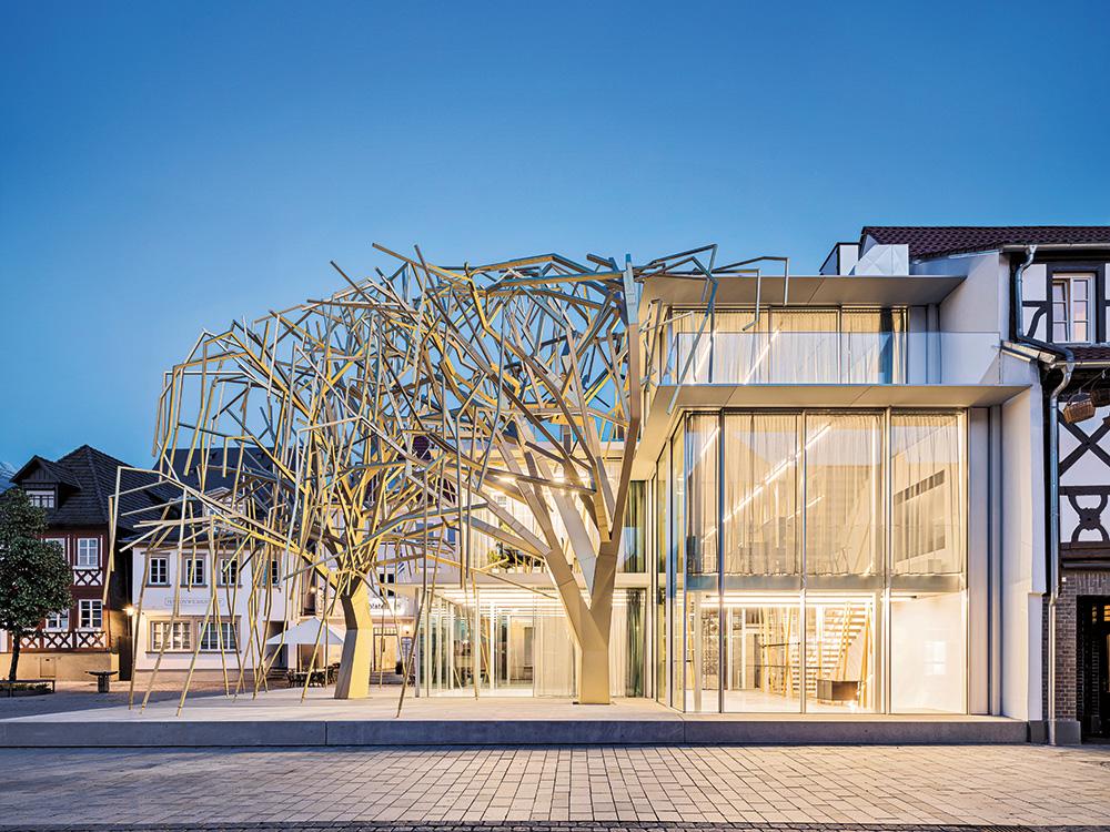 Der transparente Pavillonbau und die zwei metallisch leuchtenden Bäume zeichnen das Volumen des rückgebauten Bestandsbaus mit Walmdach nach.