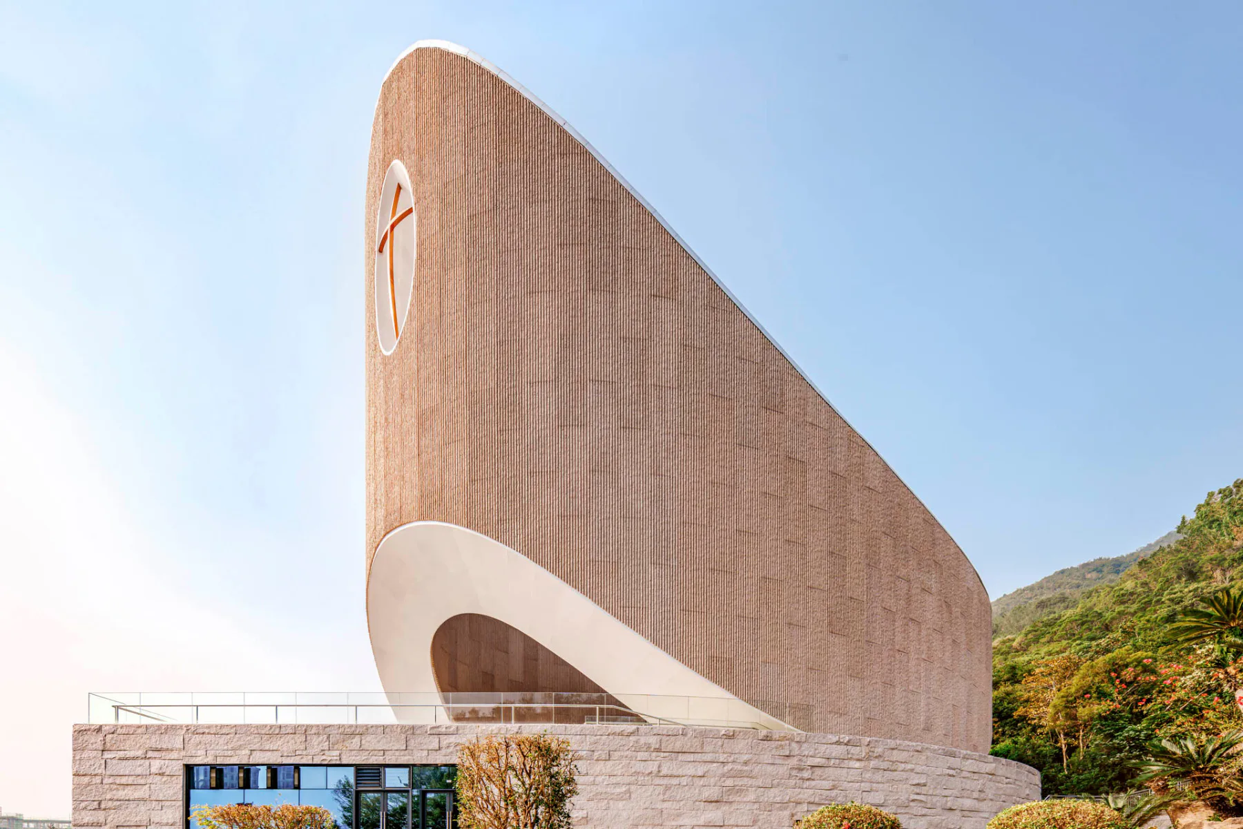 Auf der Talseite wölbt sich der ovale Kirchenbau markant empor. Der obere, geschlossene Teil der Fassaden ist mit Faserbetonelementen verkleidet.