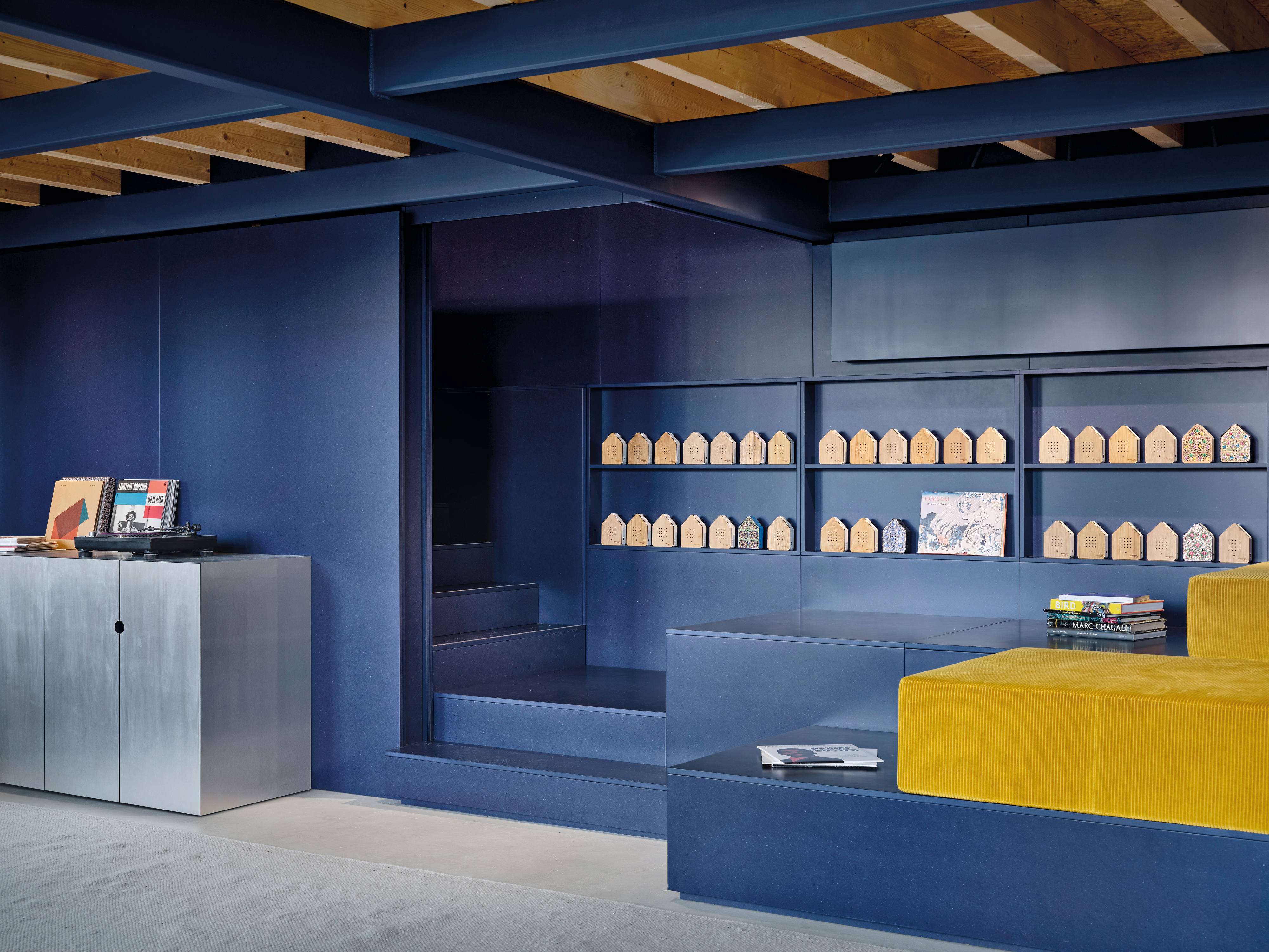 Die Lounge ist mit kobaltblau eingefärbten MDF-Platten gestaltet. Hinter einer Rückwand verbirgt sich eine Treppe, die auf die Galerie führt. Sitzstufen und Regalflächen verwandeln den Bereich in eine Lese- und Ausstellungszone.