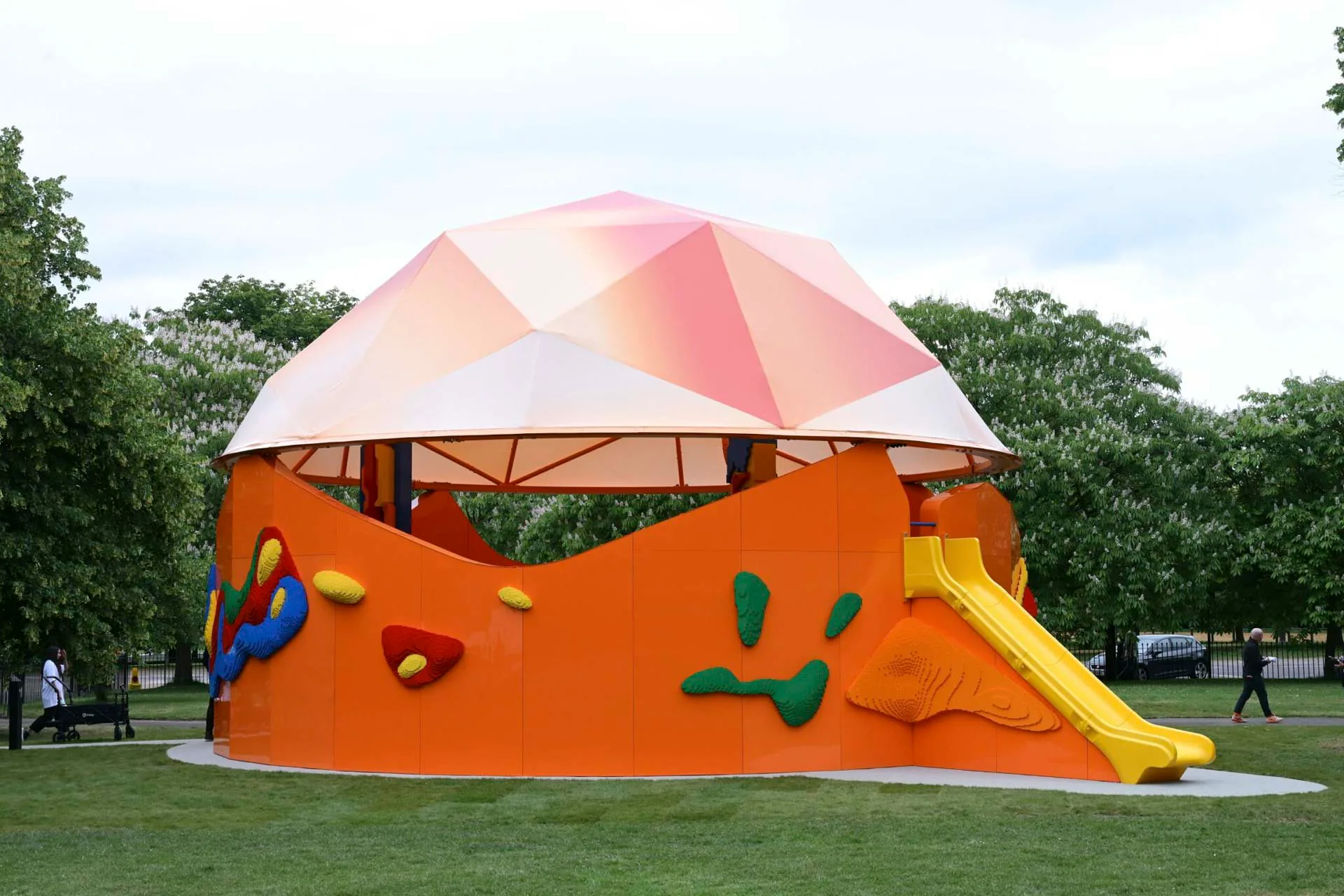 Grüne, gelbe, blaue und rote Legosteine zieren den orangefarbenen Serpentine Play Pavilion. The Play Pavilion 2025, entworfen von Sir Peter Cook (Peter Cook Studio Crablab), in Zusammenarbeit mit Serpentine und the LEGO Group.