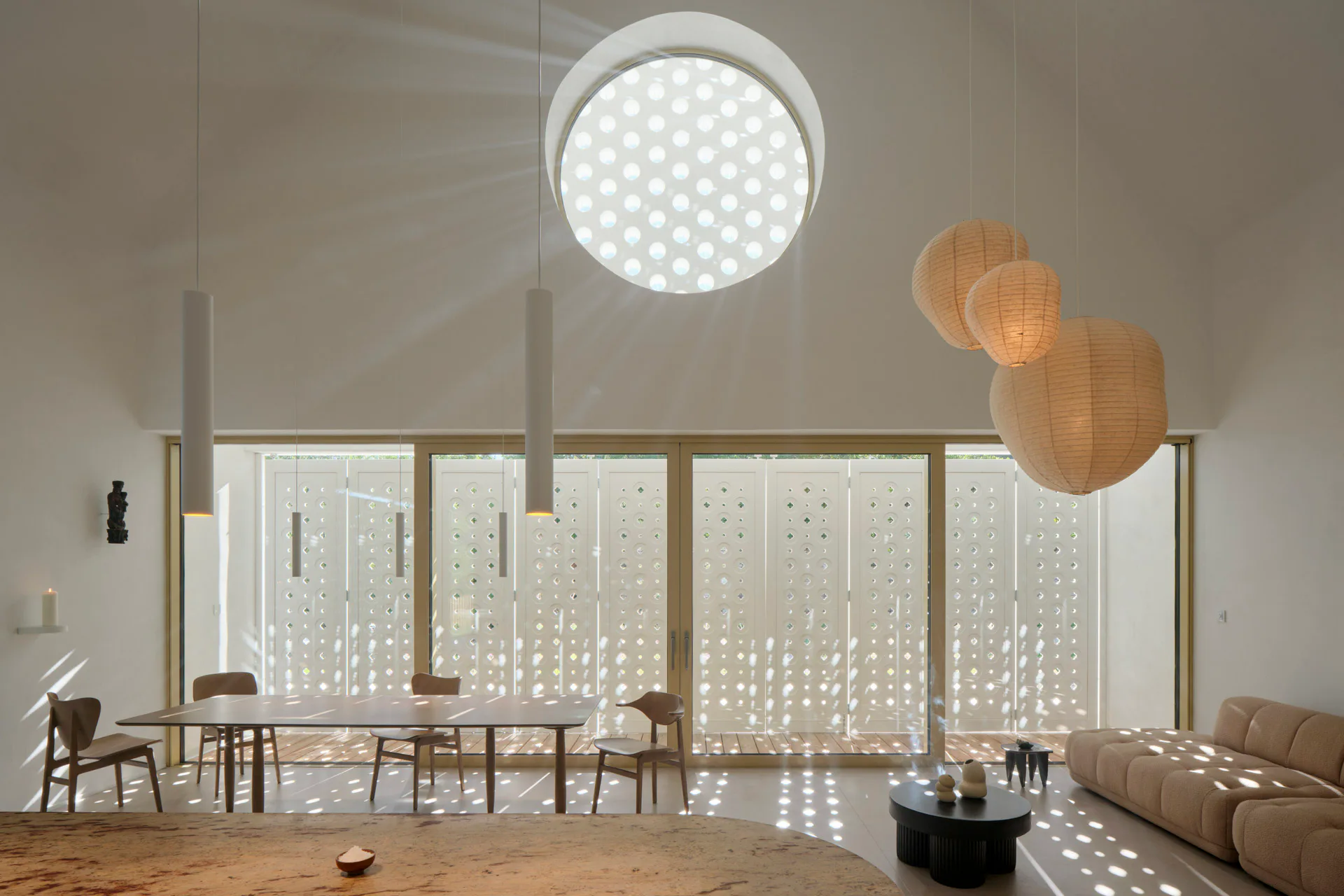 Innenraum mit großem Tisch, Sofa und Kücheninsel, geprägt von Fensterfront, rundem Giebelfenster mit perforiertem Sonnenschutz erzeugen Lichtspiel.