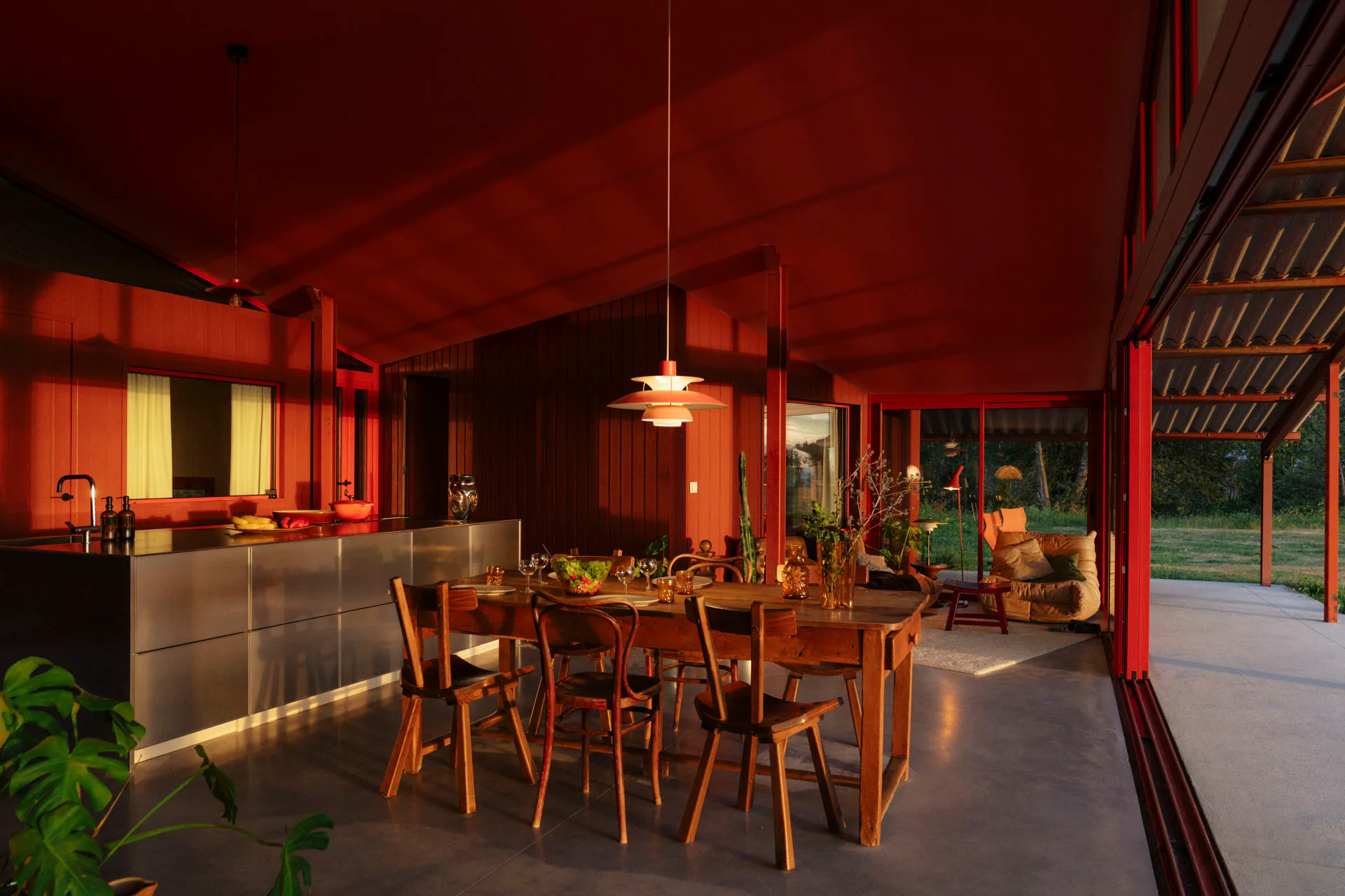Eine große Kücheninsel aus gebürstetem Edelstahl bildet das Herzstück des Raums, kontrastiert das Holz der Einbauten und reflektiert zugleich das Lichtspiel der roten Umgebung.