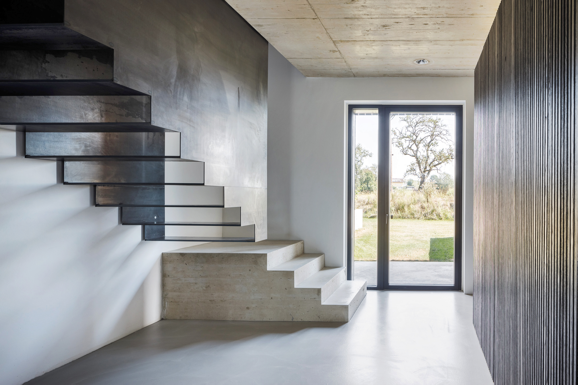 Eine filigrane Stahltreppe führt hinauf ins Obergeschoss des Wohnhauses. Ihre schwarze Farbgebung kehrt auch an der Holzhülle des Hauses wieder (rechts).
