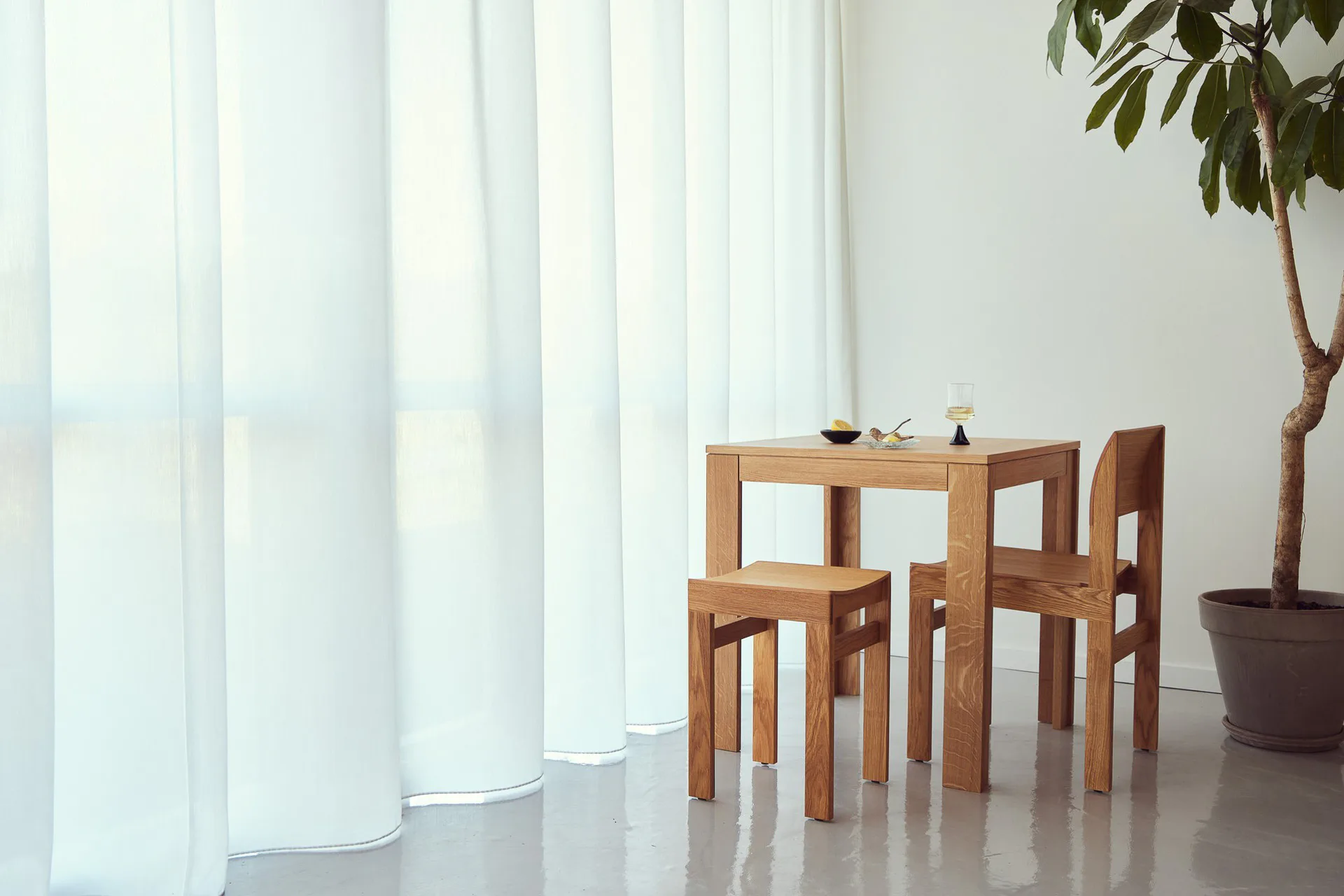 Tisch, Stuhl und Hocker aus Massivholz bilden eine kompakte Einheit mit klaren Querschnitten. Die sichtbaren Fügungen zeigen die einfache, statisch schlüssige Konstruktion.