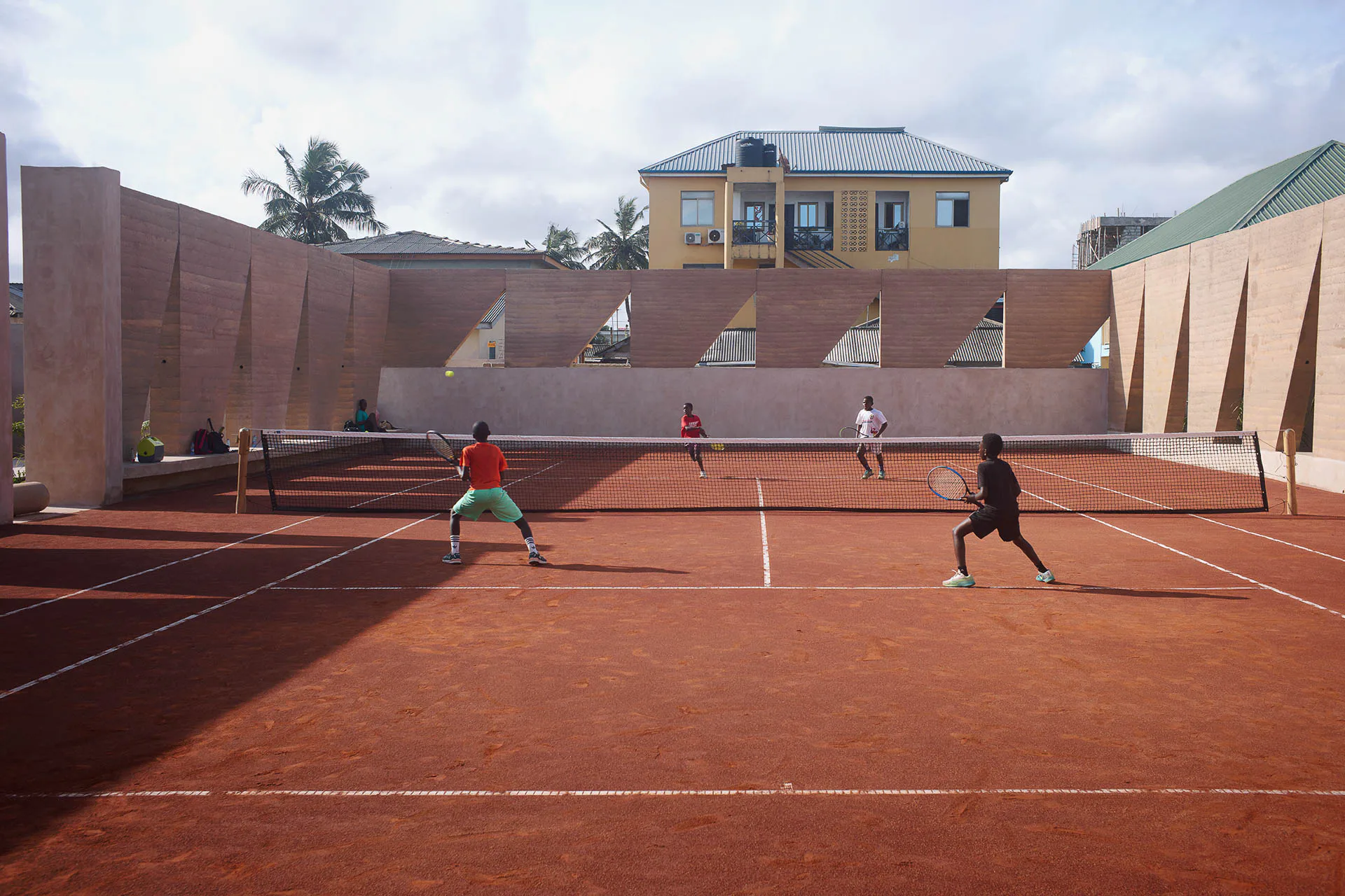 Eine 4 m hohe Stampflehm-Mauer umsäumt den Tennisplatz mit einem subtilen Rhythmus aus geschlossenen Flächen und geometrischen Einschnitten.