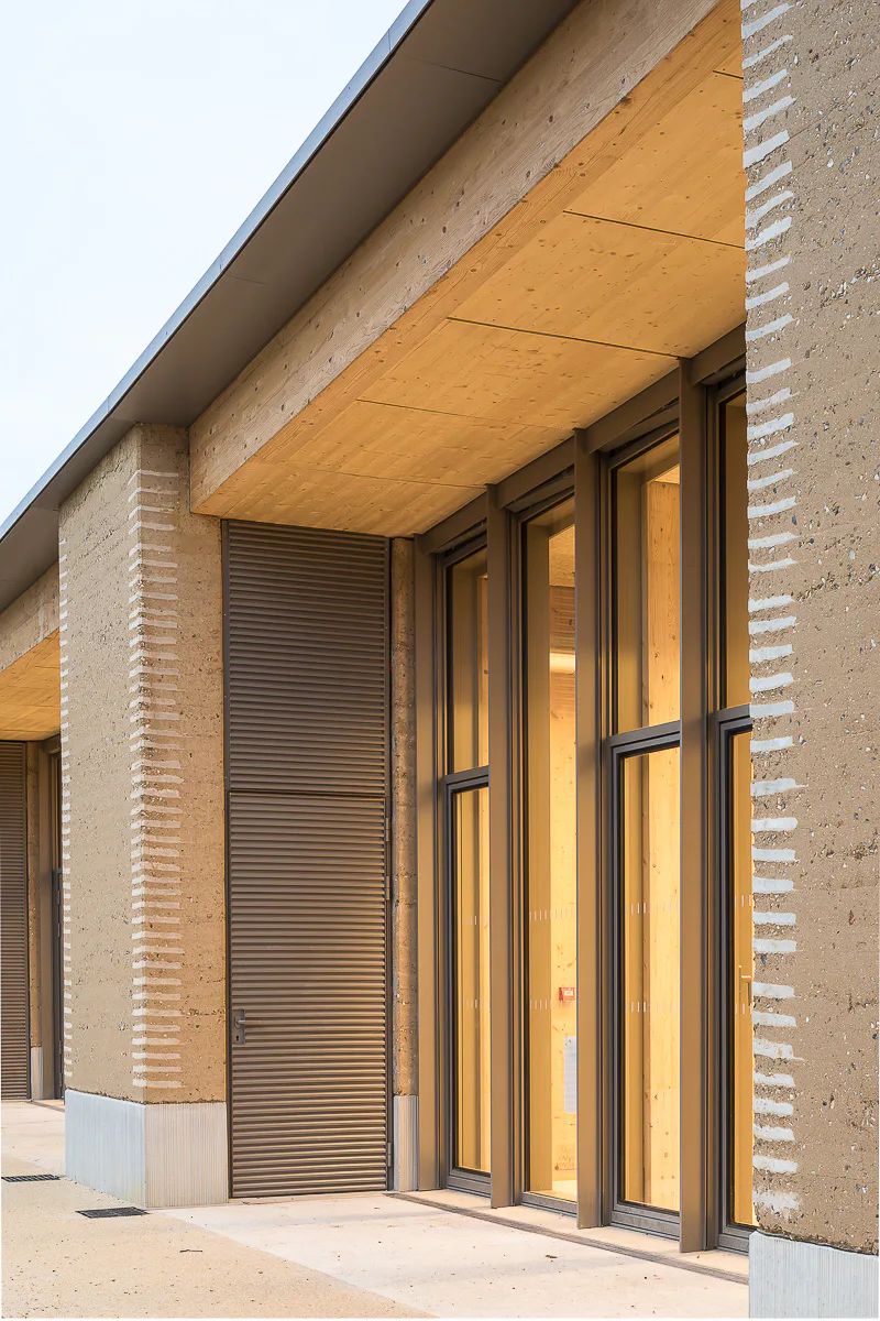 Fassade aus tragendem Stampflehm und Sockel aus kohlenstoffarmen, strukturiertem Beton