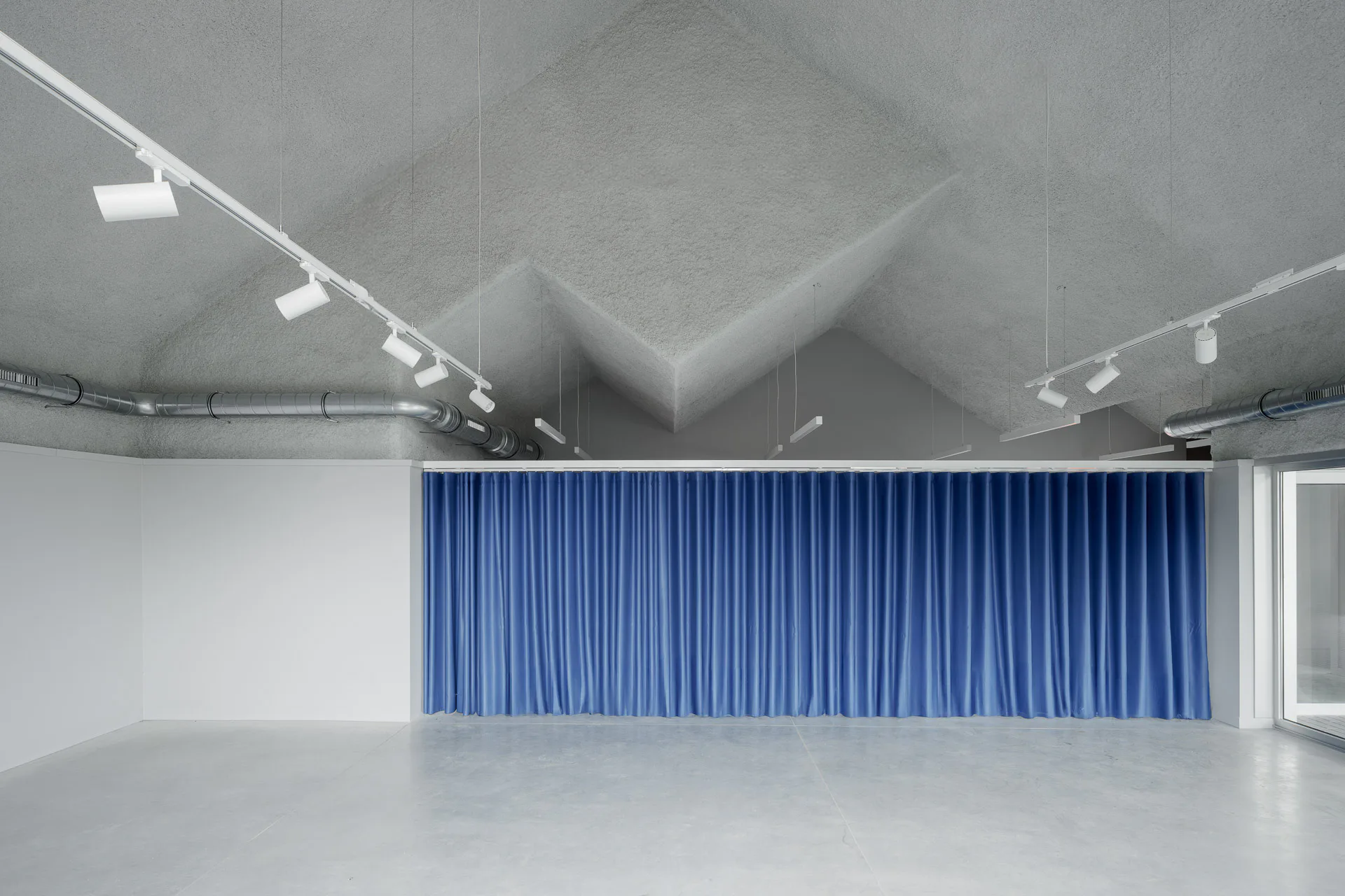 Ein blauer Vorhang trennt den Ausstellungsbereich und den Multifunktionsraum optisch voneinander. Die Faltungen des Daches stülpen sich nach innen.