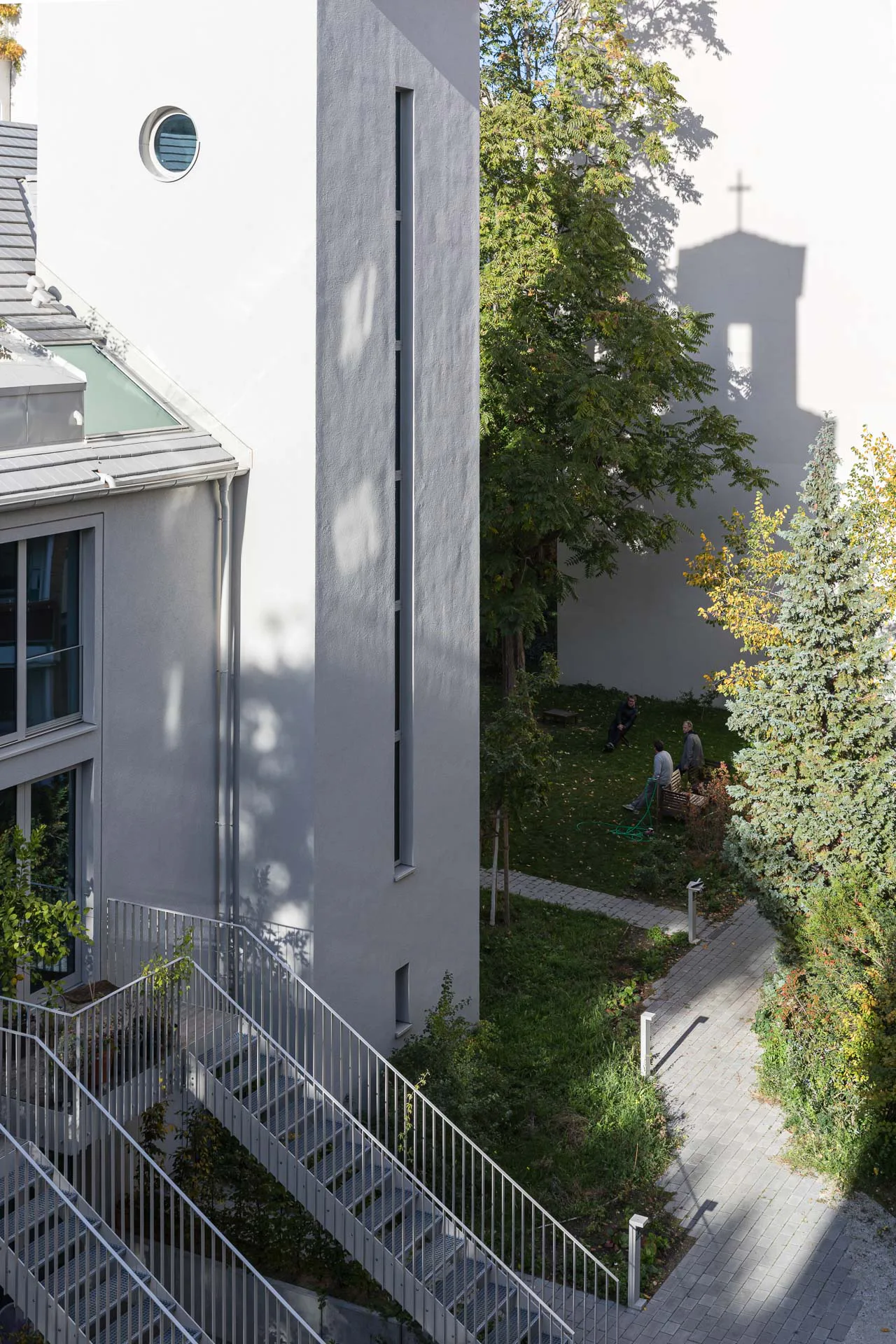 Gepflasterte Wege verbinden die Wohnanlage mit den öffentlichen Grünflächen. Ein vertikales Fensterband beleuchtet das Innere des Glockenturms.