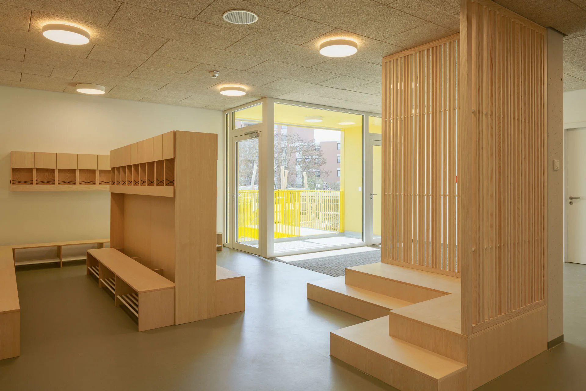 Eine Garderobe aus hellem Holz und eine Sitztribüne mit rückseitiger Lattenwand harmonieren mit dem grau-grünlichen Linoleumboden im Eingangsbereich der schulvorbereitenden Einrichtung.