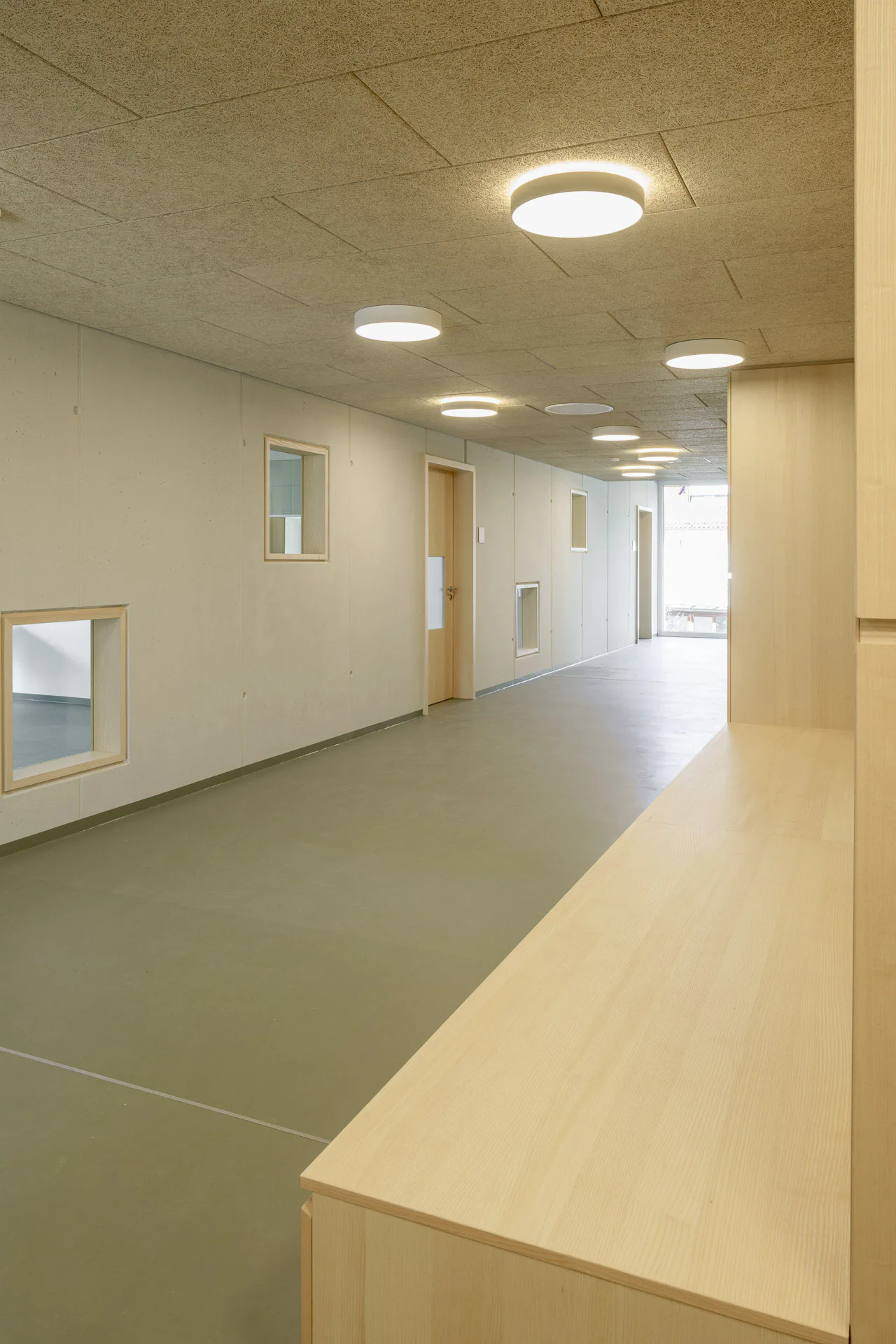 Ein großes Fenster beleuchtet den langen Flur vor den Gemeinschaftsräumen. Kleine Glasöffnungen in den Holztüren sorgen zusätzlich für natürliches Licht.