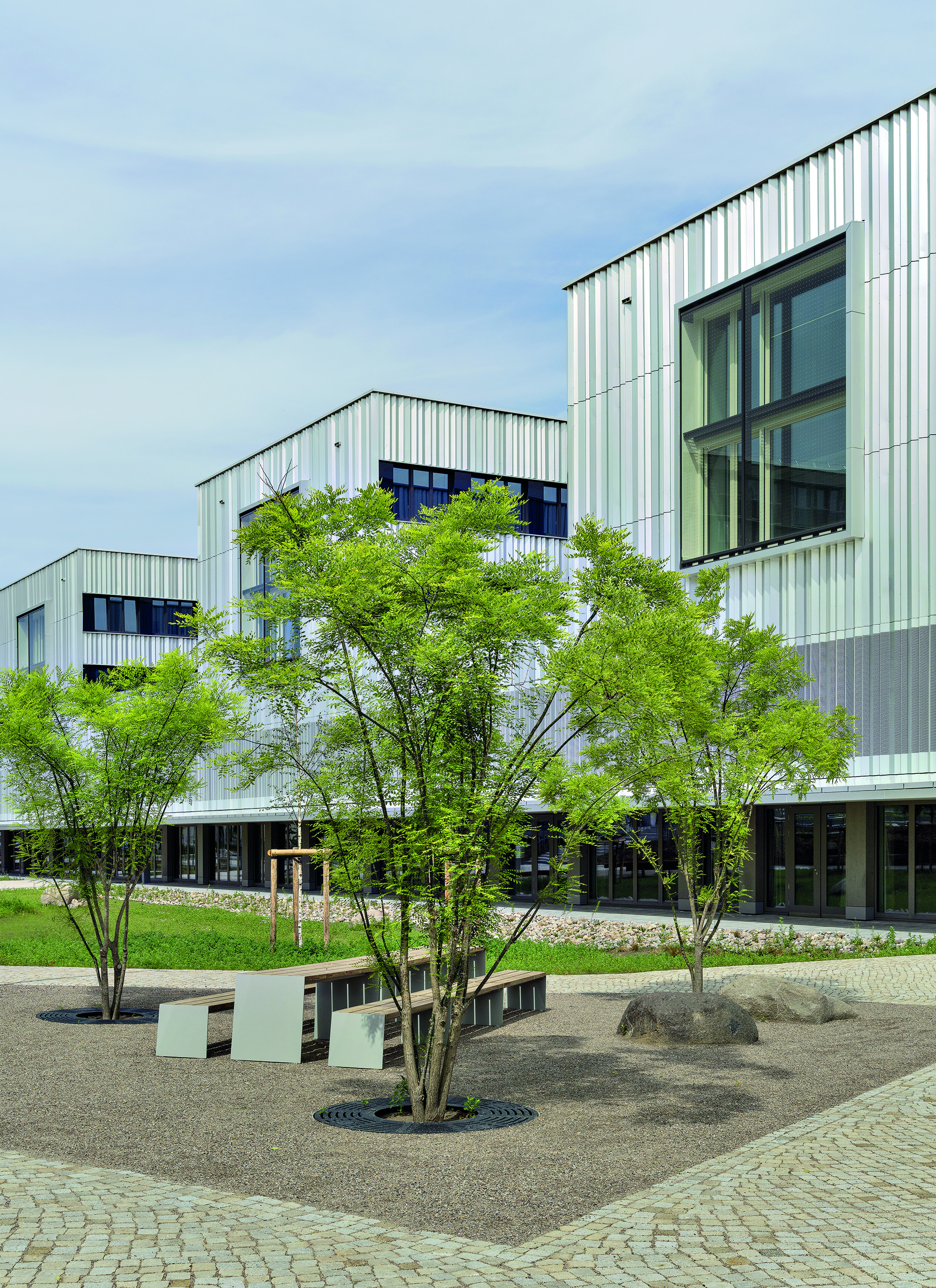 The university building is enveloped by continuous vertically folded sheet aluminium without visible horizontal fire barriers in front of the floor slabs. The two storeys tall and six metres wide southern facade box window contrasts with the serial character of the residential buildings.